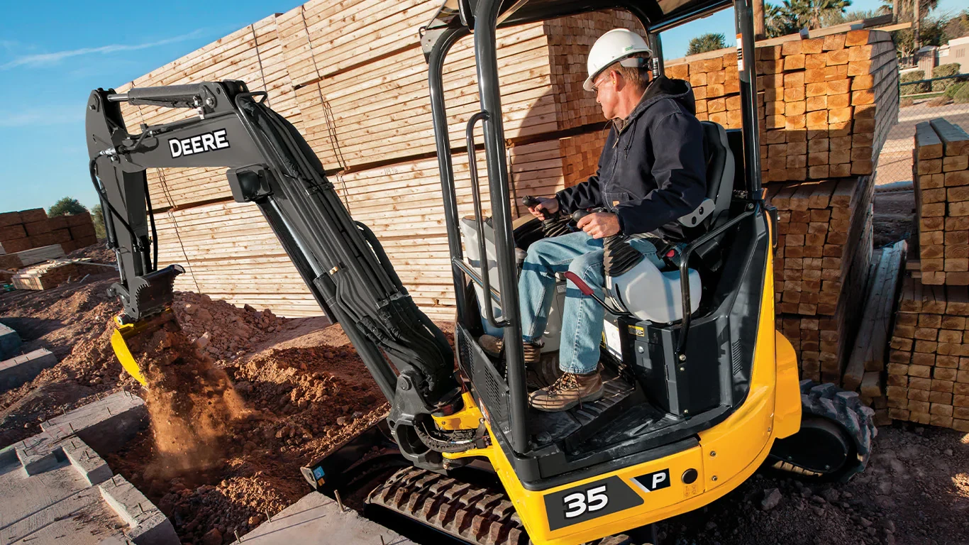 Man operating a John Deere 35P Mini Excavator that is digging in the dirt with pallets of lumber stacked behind him.