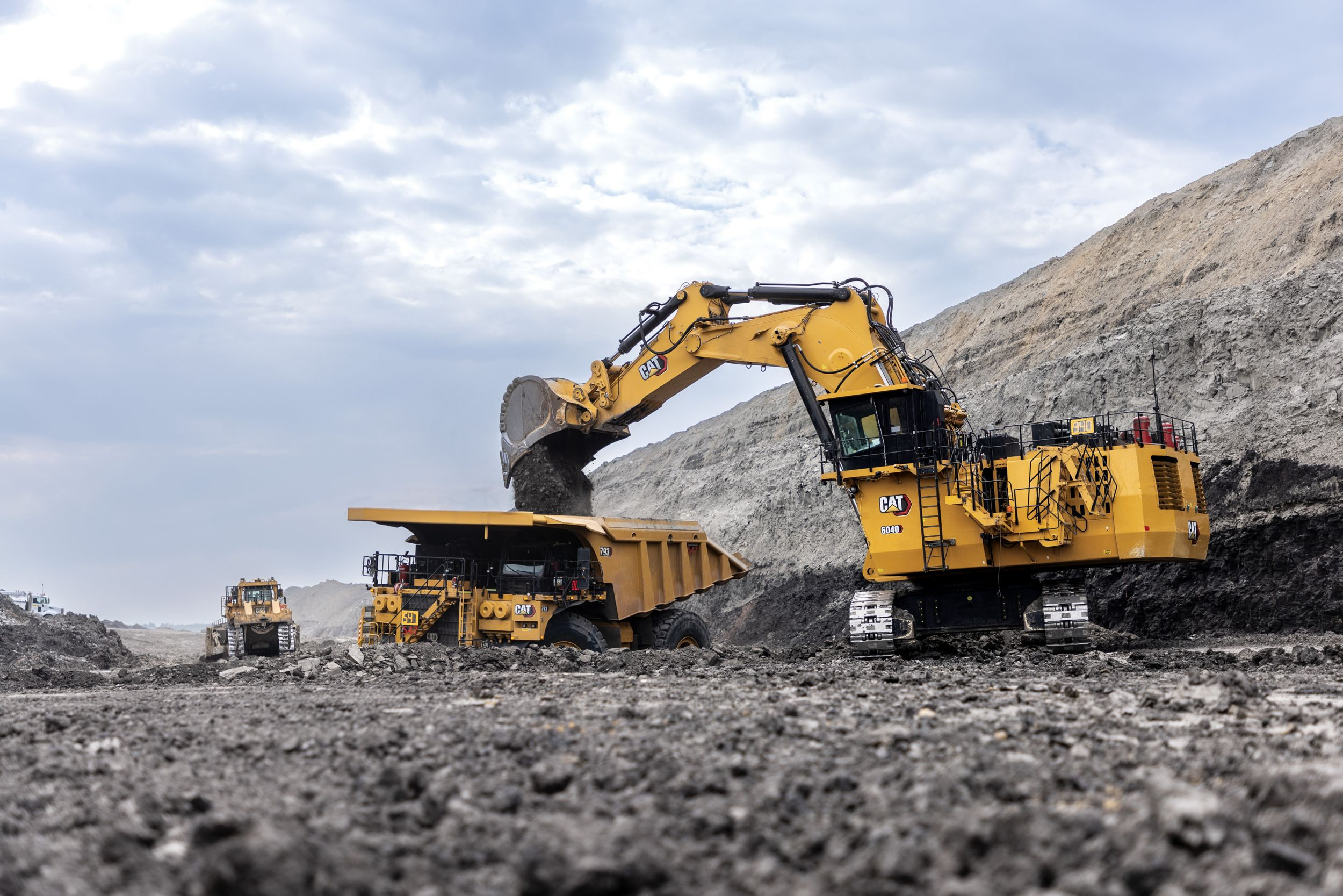 Cat 6040 mining shovel loading material into a large haul truck at an open pit mine under cloudy skies.