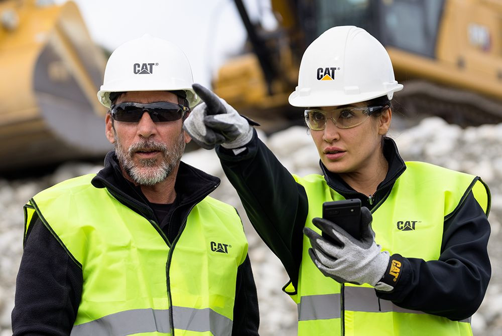 Two workers in CAT hard hats and protective gear review a phone while pointing ahead on a construction site.