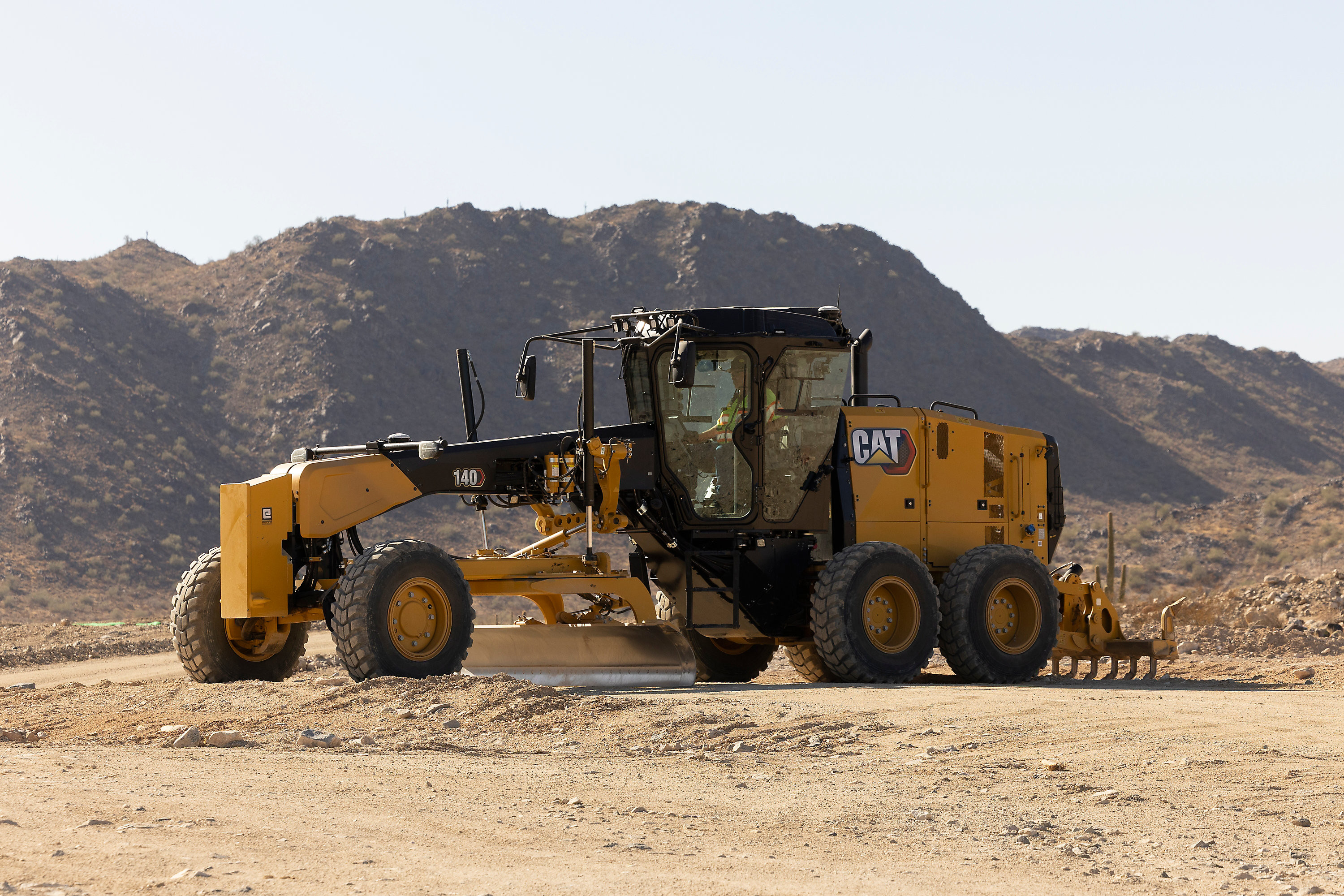 Left side view of a Cat 140 Motor Grader operating on a dirt job site with dirt piles in the background.