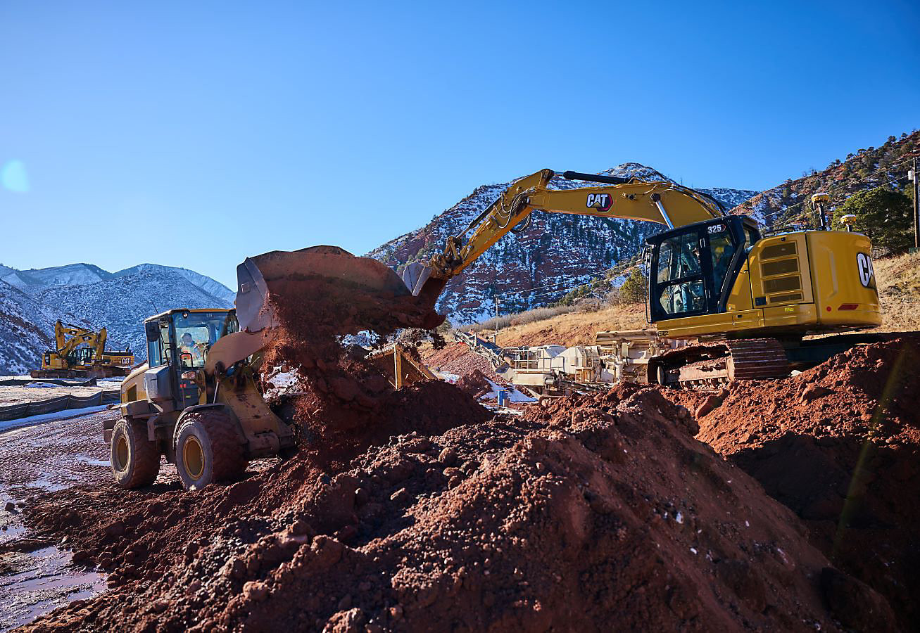 Cat 325 Excavator digging in the dirt alongside a Dozer while other heavy equipment and hills can be seen in the background.