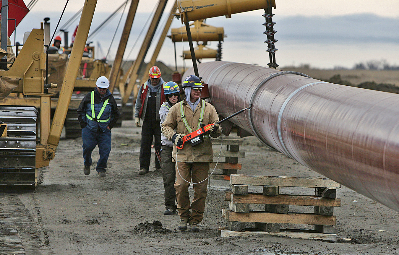 A pipeline being held by multiple pipelayers while workers jeep the pipe, cribbing is seen on the ground.
