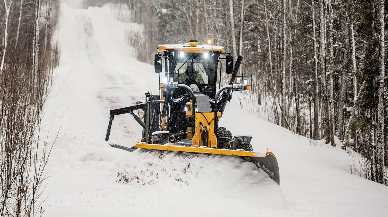 Man operating a John Deere that is plowing snow on a sloped path with trees on each side.