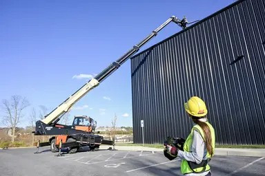 Worker in safety gear operates a JLG Telehandler with an extended boom reaching the top of a tall metal-sided building.