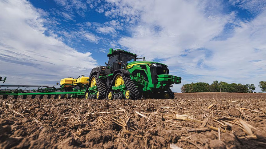 Low angle view of a green John Deere 8-series tractor planting in a field with dramatic sky and textured soil foreground.