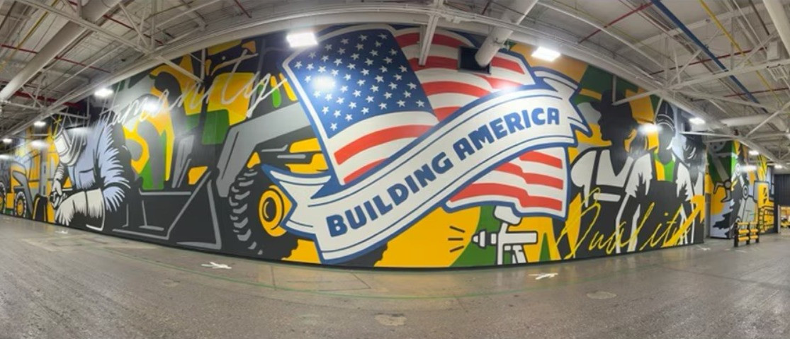 Large factory mural featuring workers, machinery, and an American flag banner with text "Building America."