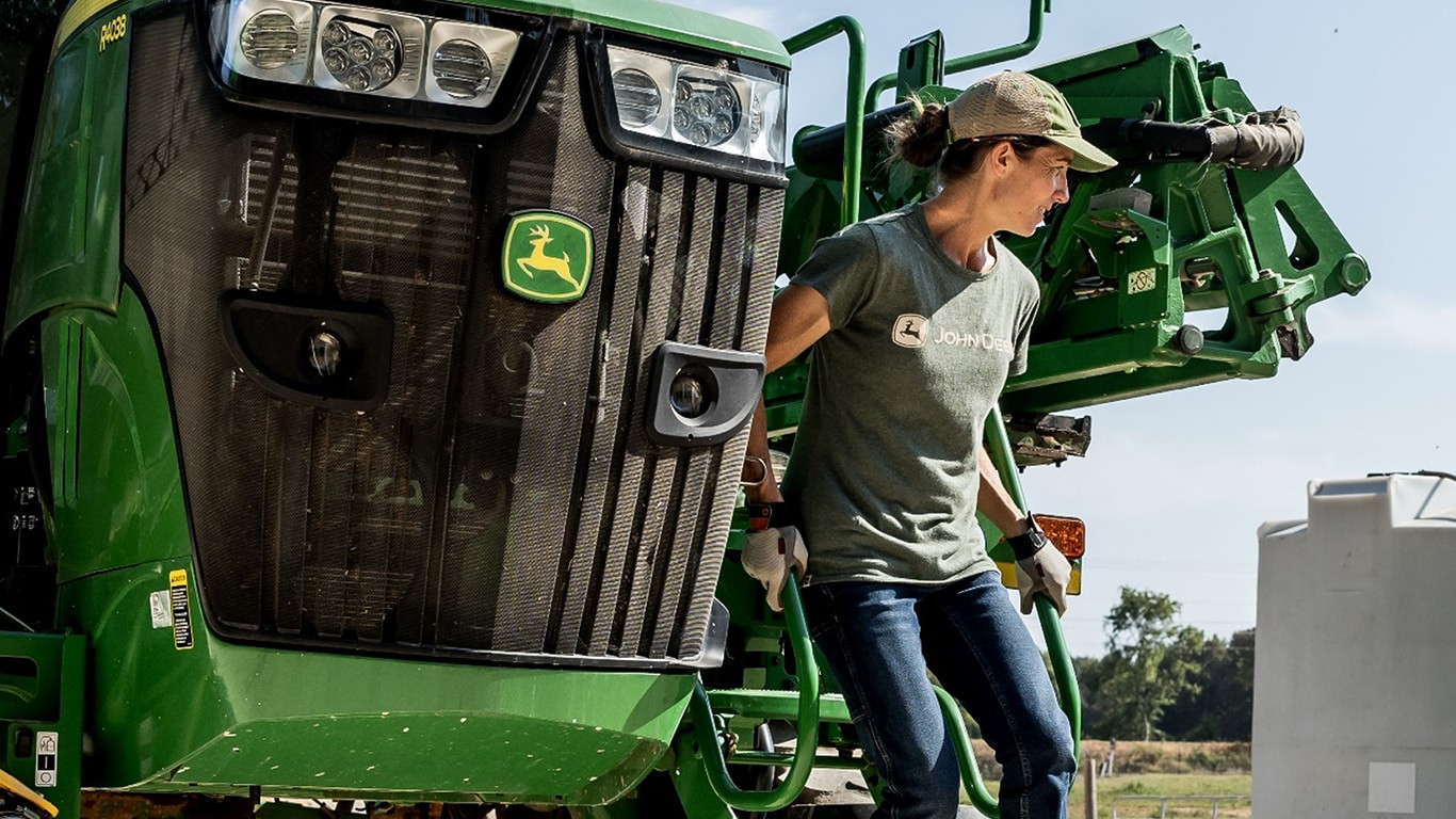 A woman is descending the steps of a John Deere combine and is wearing a grey john deere tshirt and jeans.