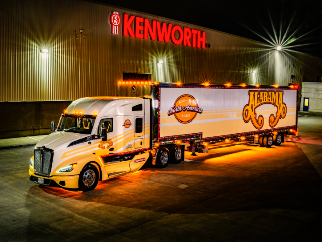 Left side view of a white Kenworth semi truck with lights on and the text "Alabama"  and "Roll On North America Tour."