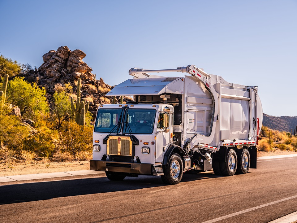 Left side view of a white Kenworth Refuse truck on the highway next to rocks and cactuses.