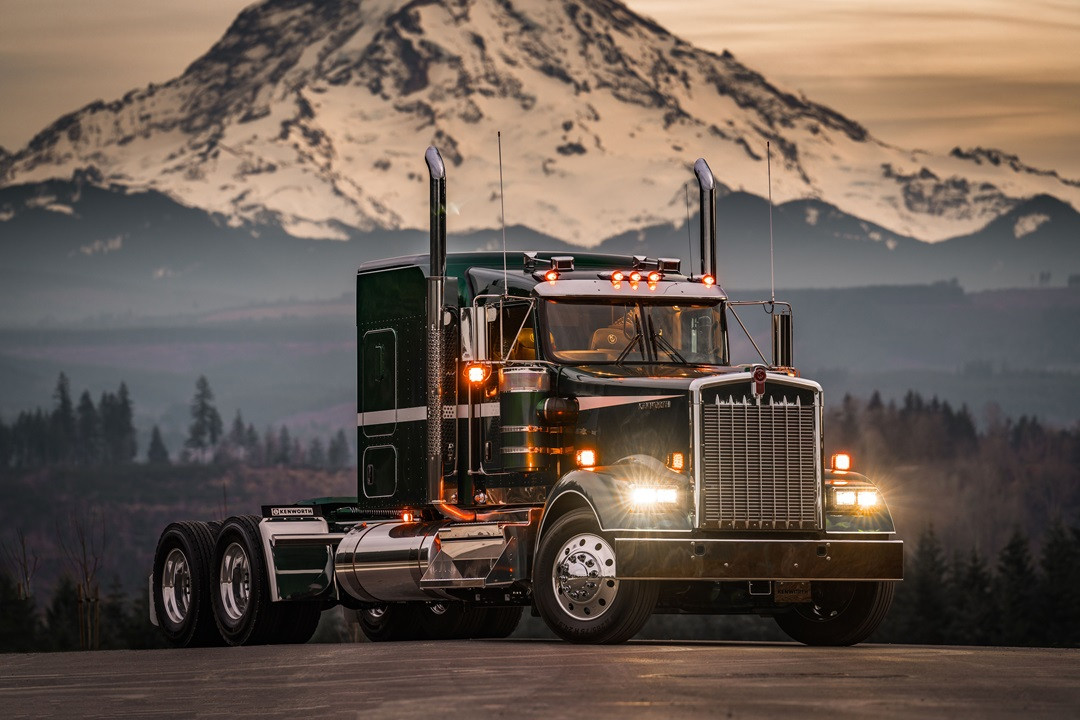 Right side view of a black Kenworth W900 Legacy semi-truck parked with headlights illuminated and a mountain in the distance.
