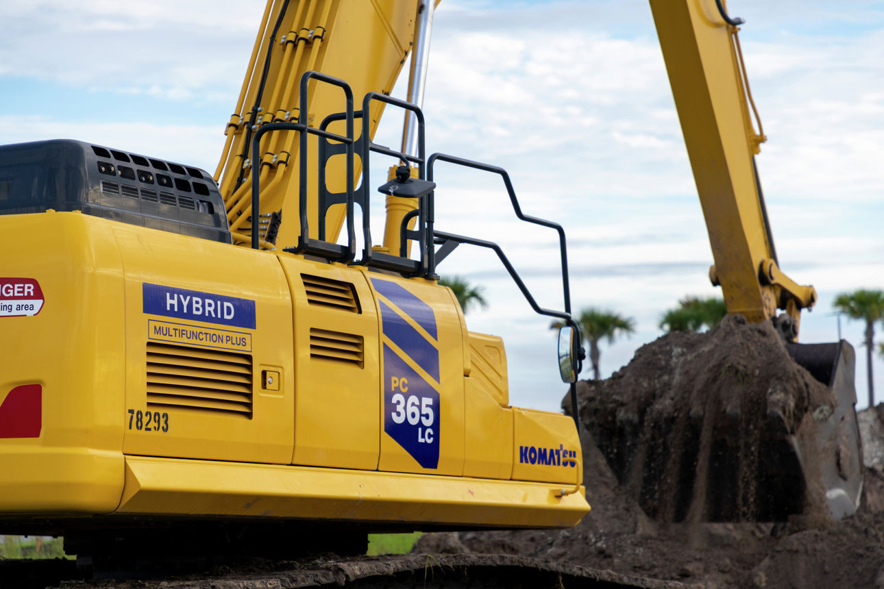 Komatsu PC365LC-11 Hybrid excavator digging soil with palm trees and cloudy sky in the background.