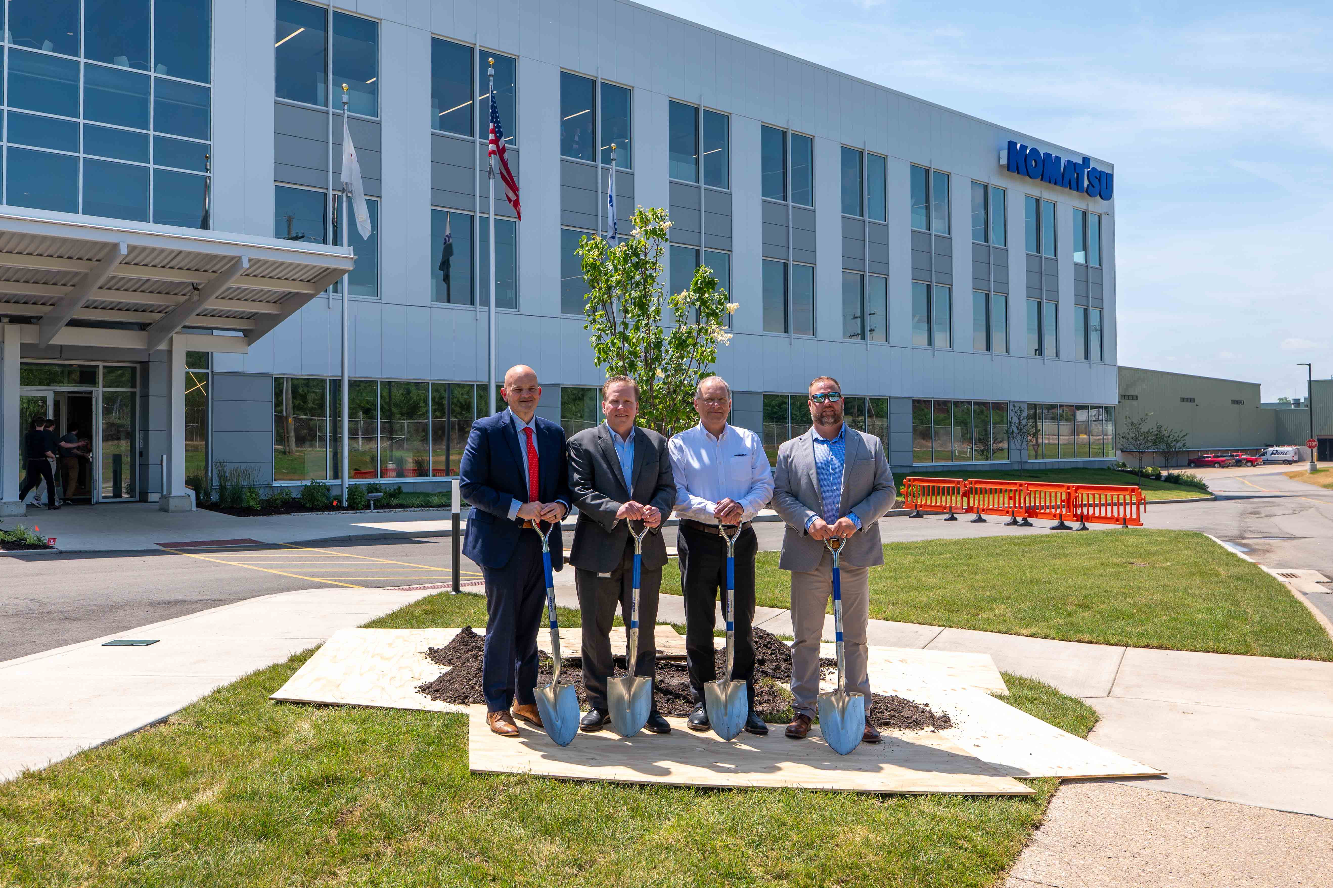 Four men pose with shovels beside a newly planted tree outside of Komatsu's Peoria facility.