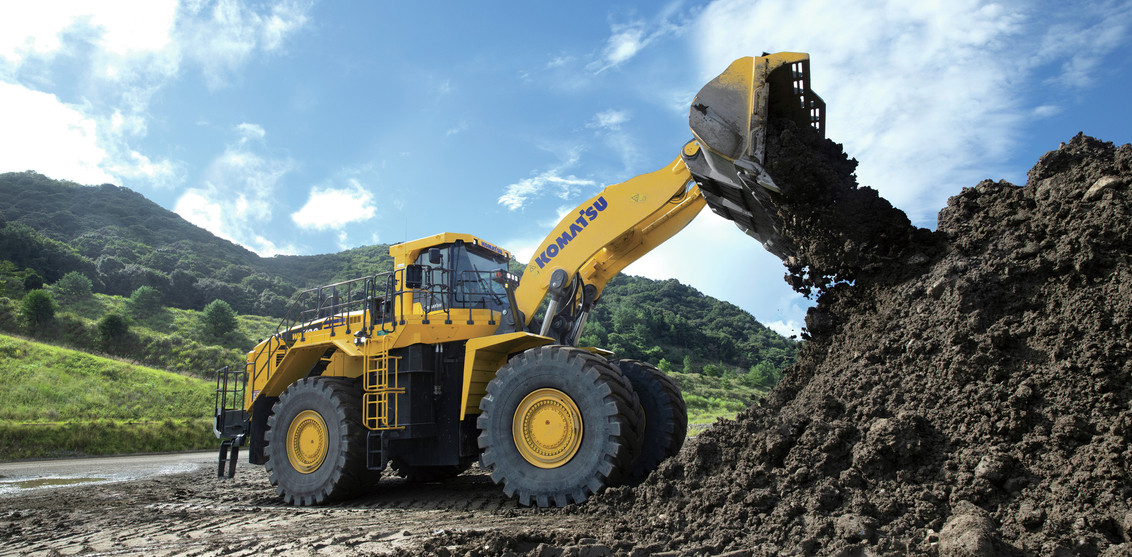 Right side view of a Komatsu WA700-8 Loader pouring the contents of its bucket into a large dirt pile with trees surrounding.