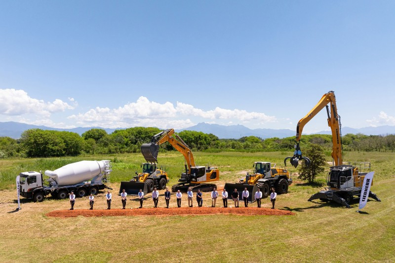 A line of men digging their shovels into the ground at a Liebherr groundbreaking ceremony with equipment parked behind them.