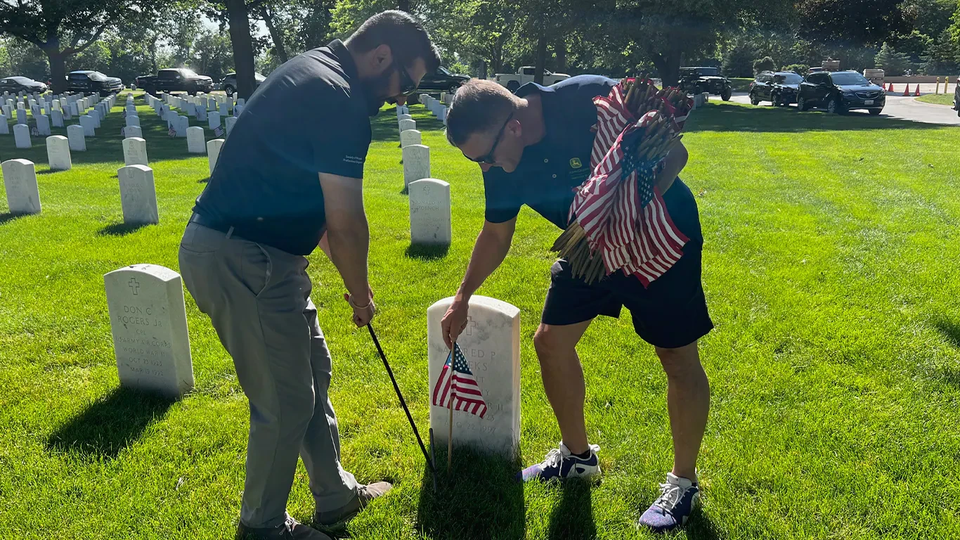 Two men placing flags in the ground by gravestones in a cemetery.