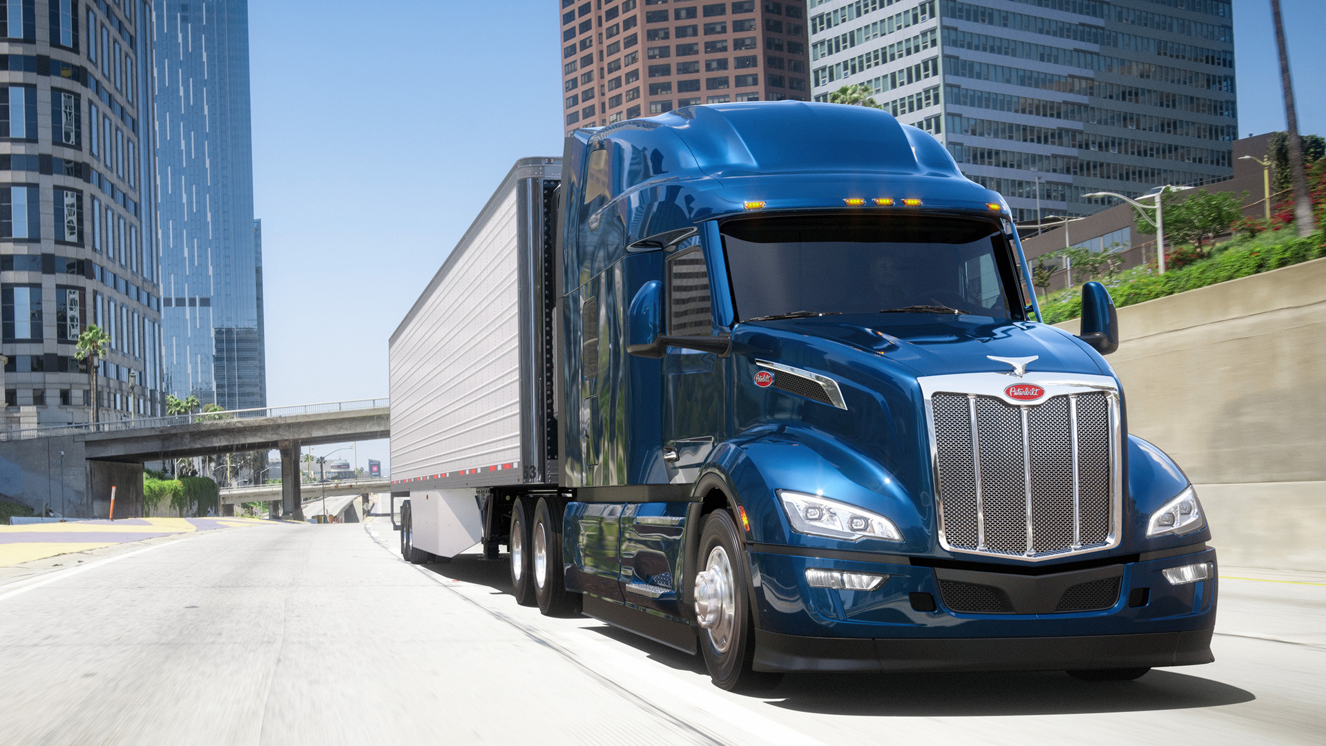 Dark blue Peterbilt semi-truck and trailer driving on a highway with high rise buildings in the background.
