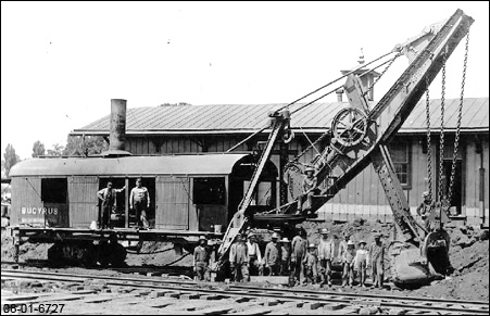 Vintage black and white Bucyrus excavator parked while workers stand on and around it, building and train tracks visible.