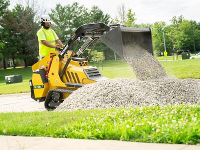 Man operating a Vermeer ML Series Mini Loader that is dumping gravel onto a pile near grass