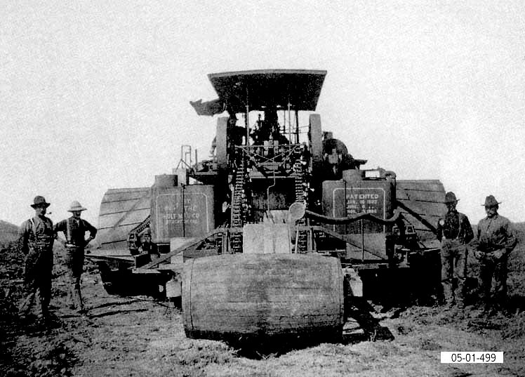 Vintage black and white early track-type tractor invention by Benjamin Holt with men standing on each side of the machine.