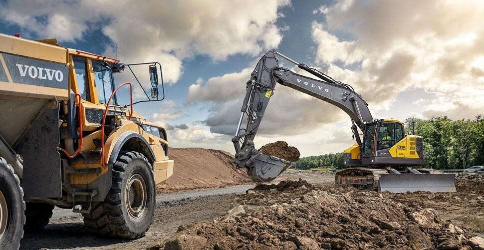 Volvo ECR355 short swing excavator digging soil beside a Volvo dump truck at a construction site under a cloudy sky.