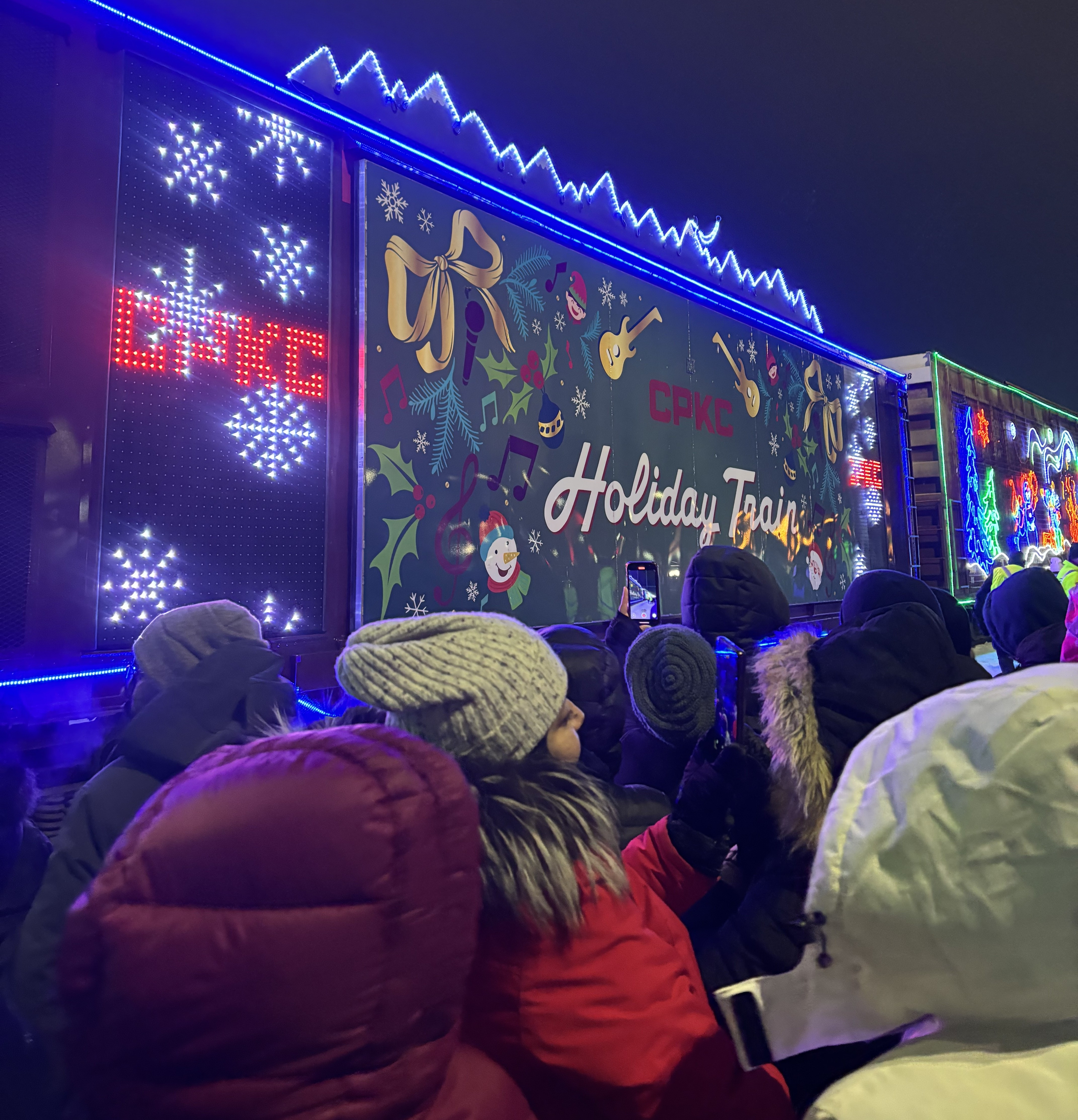 A train with bright lights and Christmas icons surrounded by people dressed warmly with text "CPKC" and "Holiday Train."